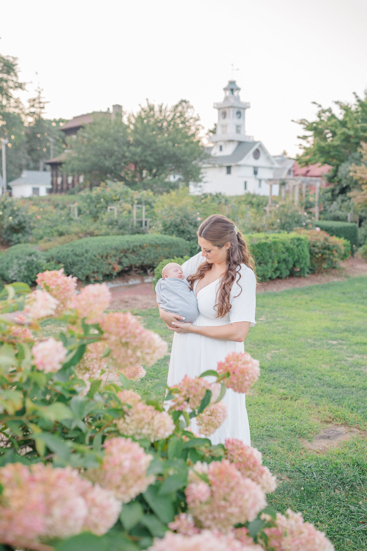 Mother lovingly holding her newborn baby in her arms, standing in a beautiful morning garden. Soft sunlight filters through the lush greenery, creating a peaceful and intimate moment between mother and child