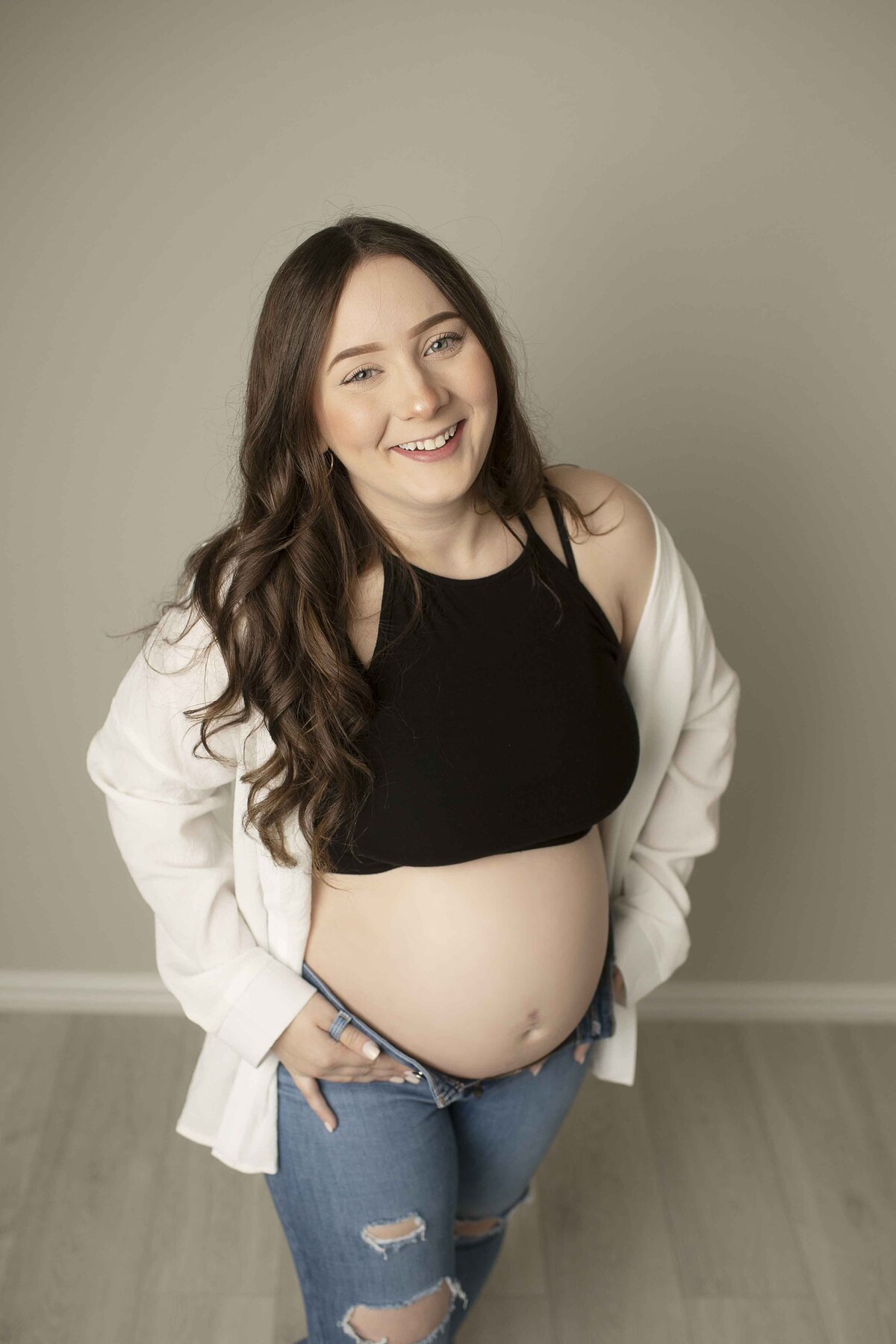 A casual and intimate maternity photo of the expectant mom in jeans and a tank top, gently cradling her belly while standing confidently in the studio, captured by Chunky Monkey Photography.