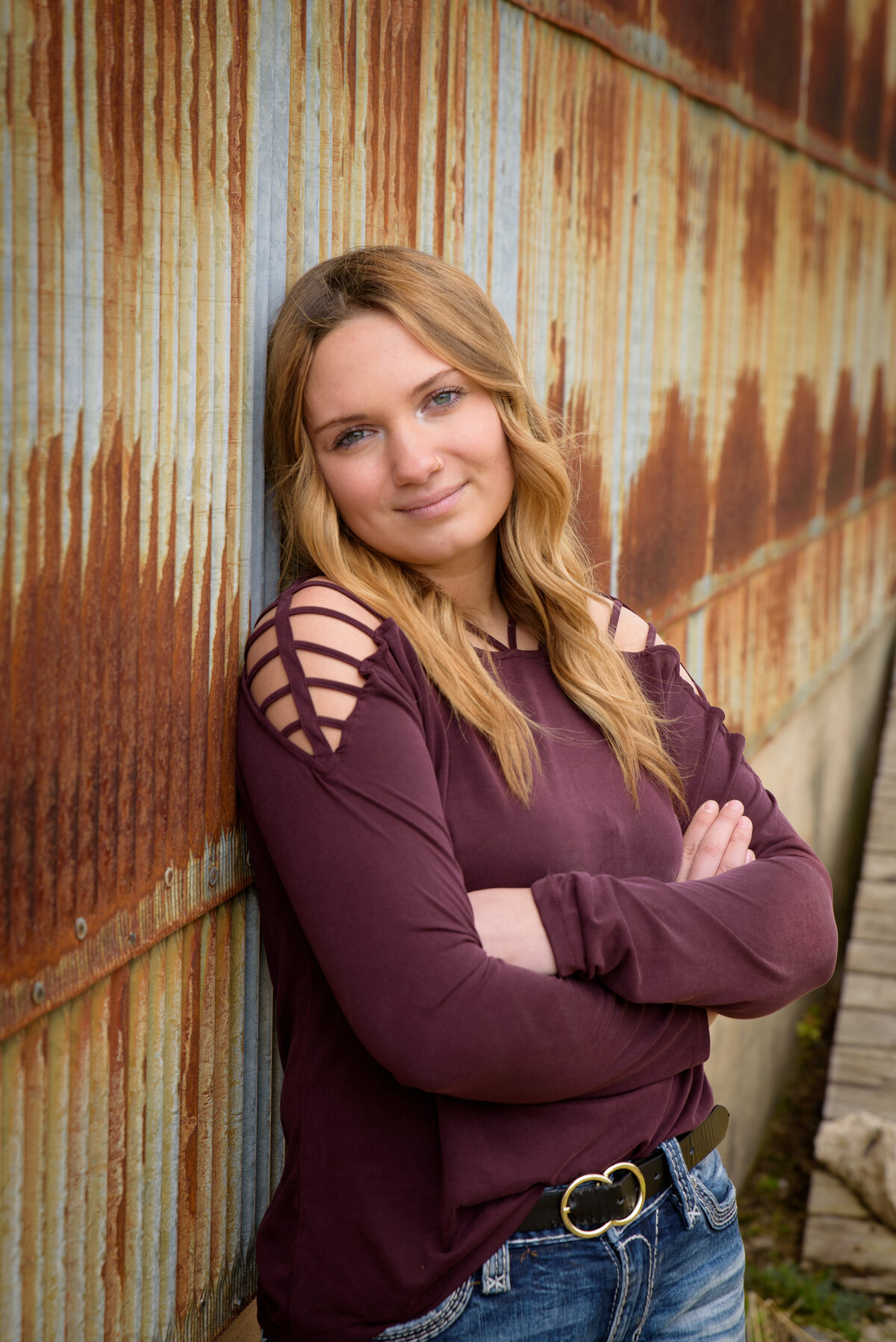 a high school senior girl is leaning against rusty corrugated metal building wearing a maroon shirt and jeans.