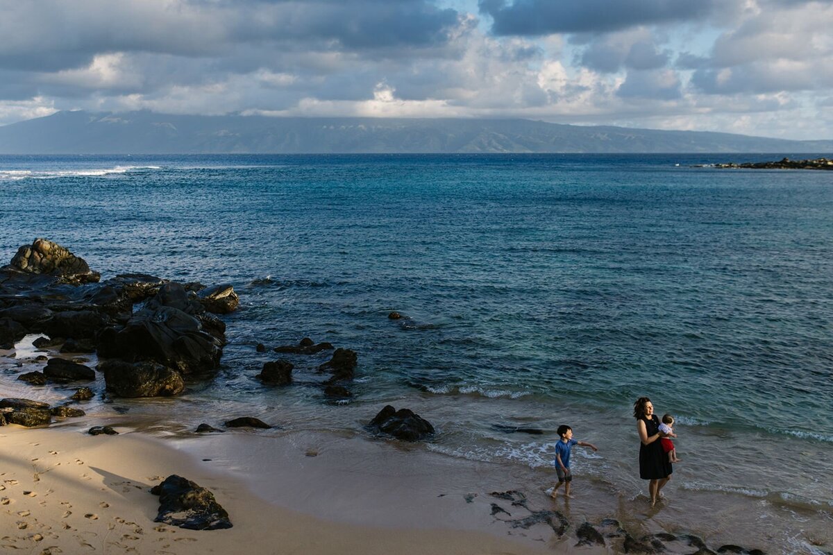 A woman holds her baby and her son follows behind as they play along the beach of Maui.