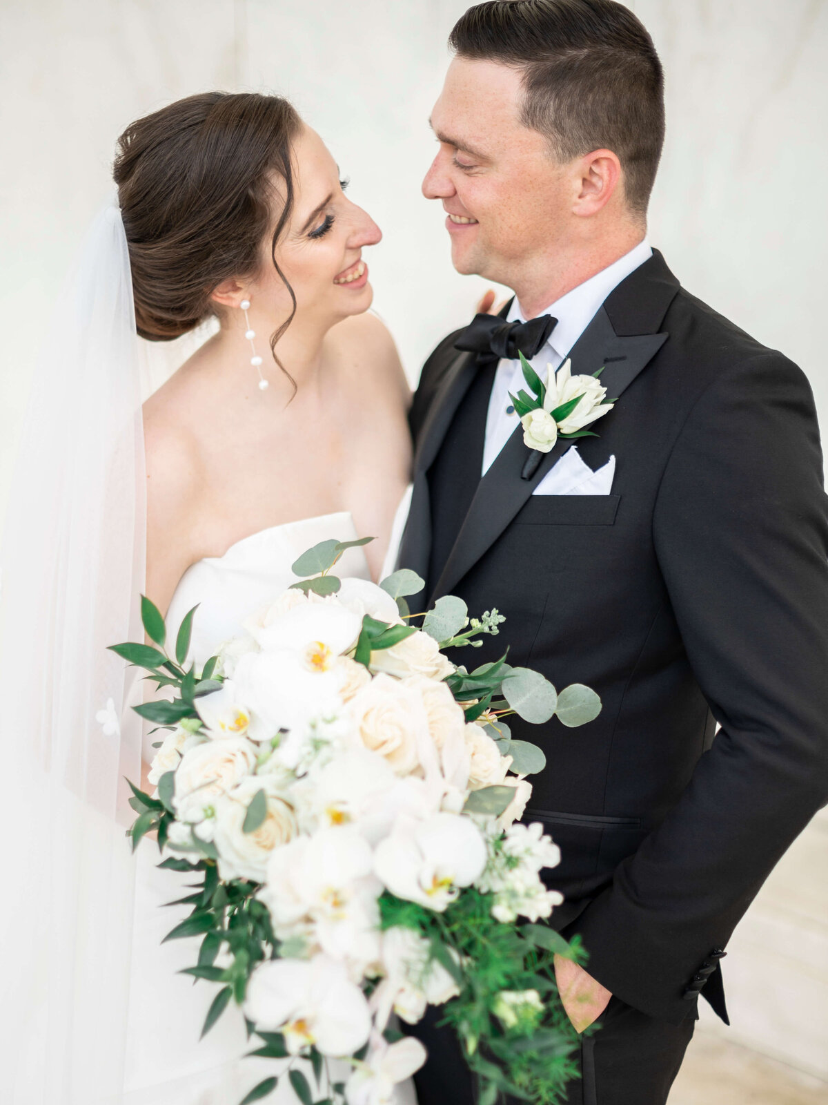 Bride and groom smiling at each other, dressed in formal attire. The bride holds a bouquet of white flowers with greenery. The groom wears a black tuxedo with a boutonniere matching the bouquet. The background is softly blurred, focusing on the couple.