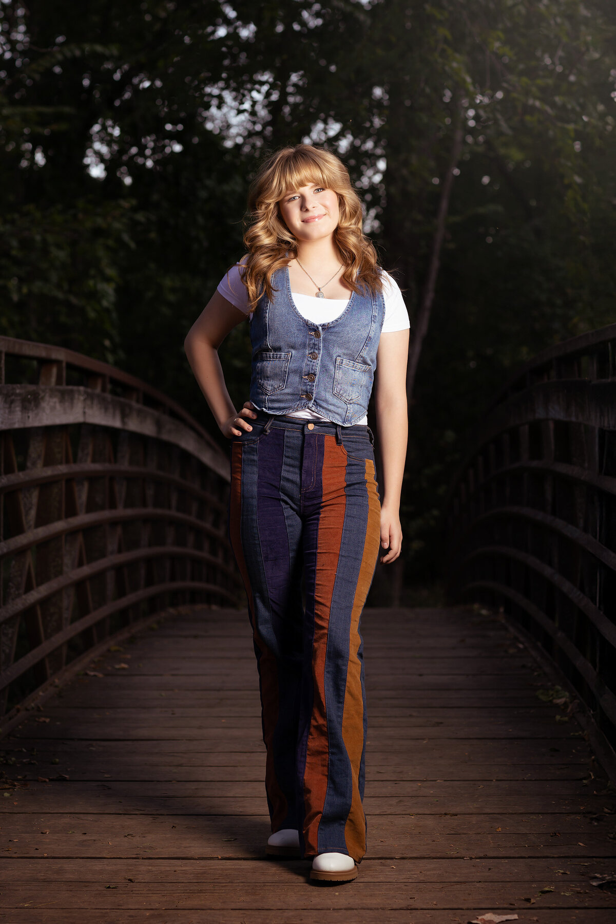 Dallas senior photography of a young woman in a denim vest and striped pants, standing on a bridge in nature.