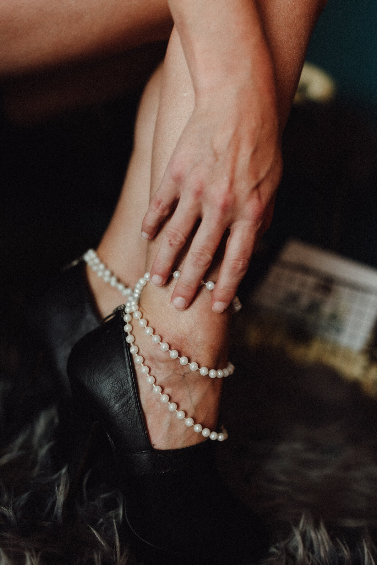 Close-up of woman’s ankle wrapped in pearls with black high heel and hand resting gently on her leg during a boudoir session.