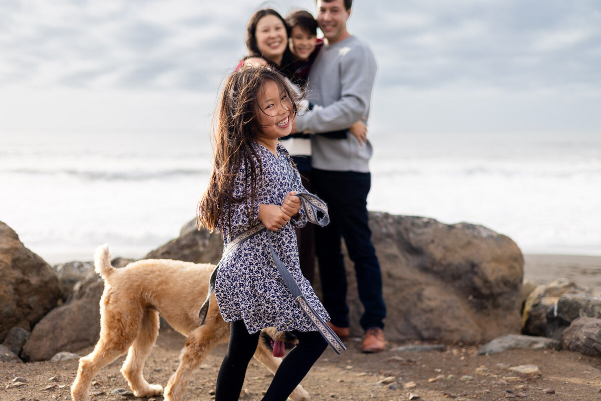 Young girl running joyfully with her dog as family smiles in the background at a San Mateo beach – Bay Area Family Portfolio – Ellobelle Photography