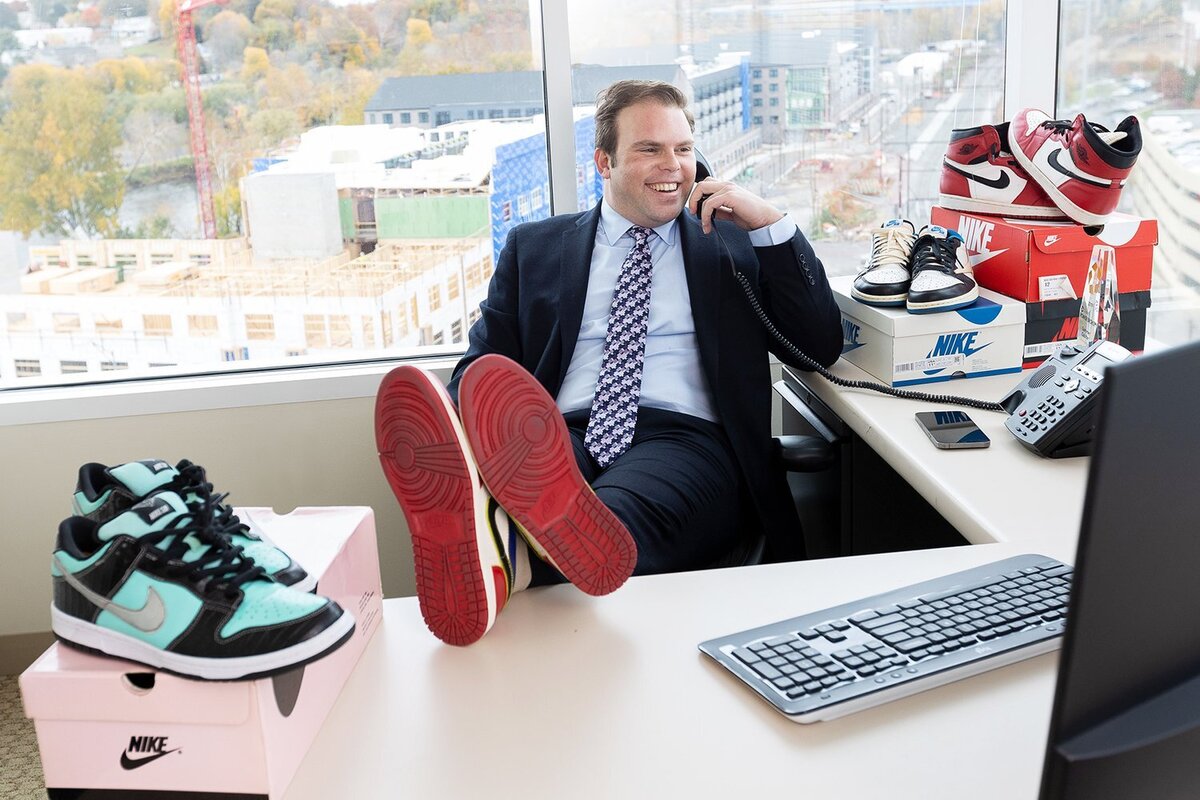 middle aged guy with his feet up on his desk, talking on the phone. There are expensive sneakers stacked on his desk around him.