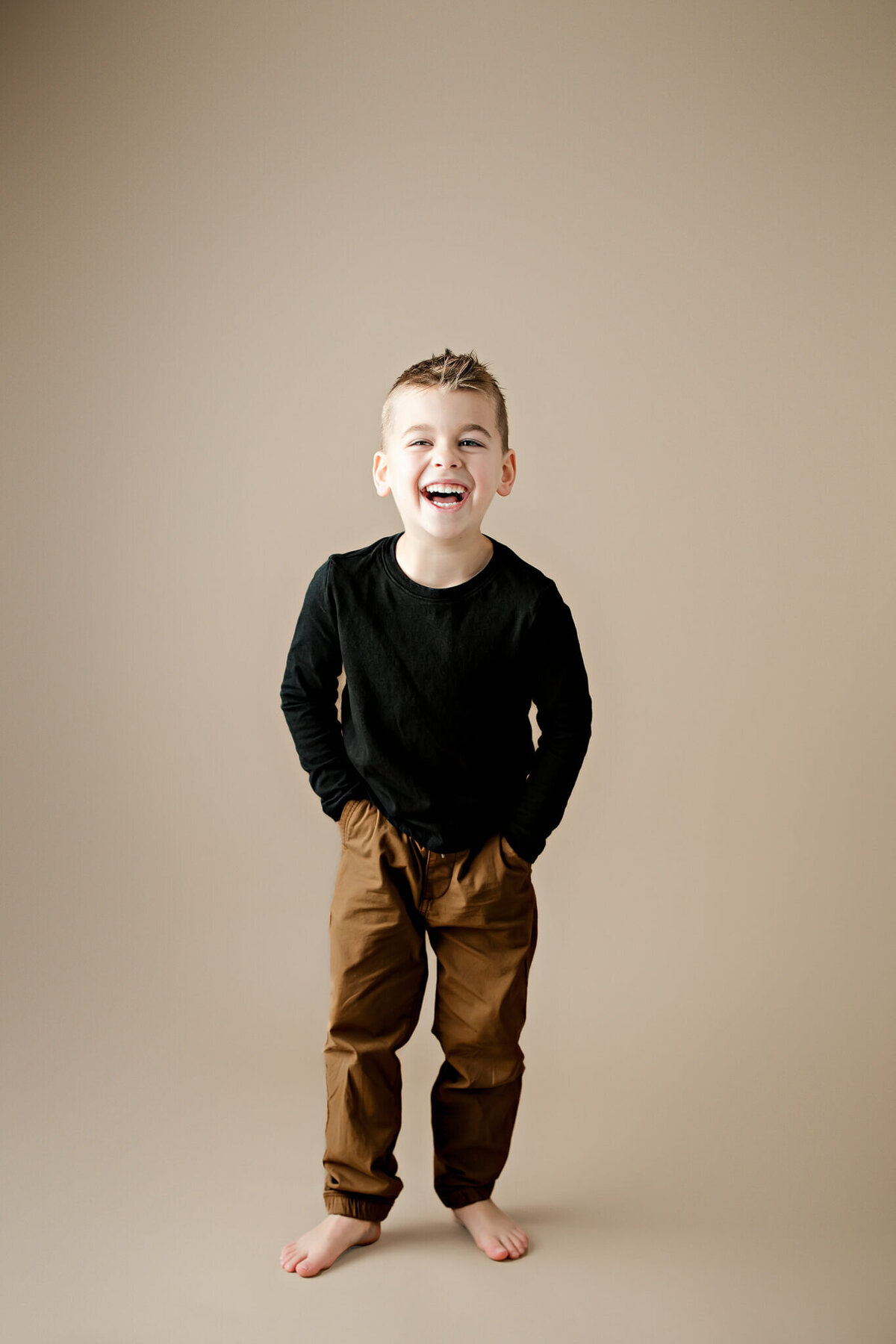 Smiling young boy with short hair, barefoot and wearing a black long-sleeve shirt and brown pants, stands against a beige background with his hands in his pockets—a perfect milestone celebration moment for any Bloomington cake smash photographer to capture.
