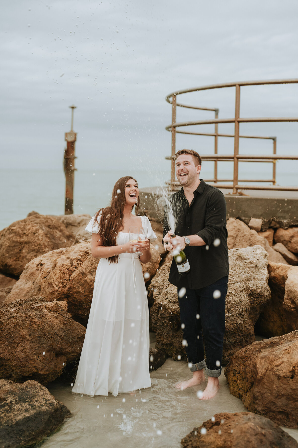couple popping champagne on the beach laughing at each other during engagement session