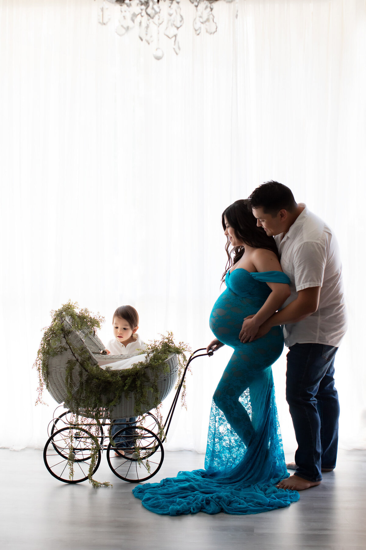 Darling family maternity photograph of a family. Dad is standing close to mom and looking over her shoulder. Mom is in a beautiful sheer lace teal gown gazing down at an empty vintage stroller and a young big brother is looking expectantly into the stroller.