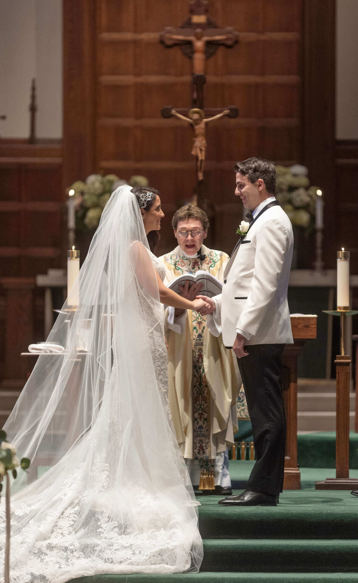 A bride and groom stand at the altar, holding hands and exchanging vows in a church decorated with white flowers. The priest stands behind them, smiling. The bride wears a long veil and gown, and the groom is in a white tuxedo.