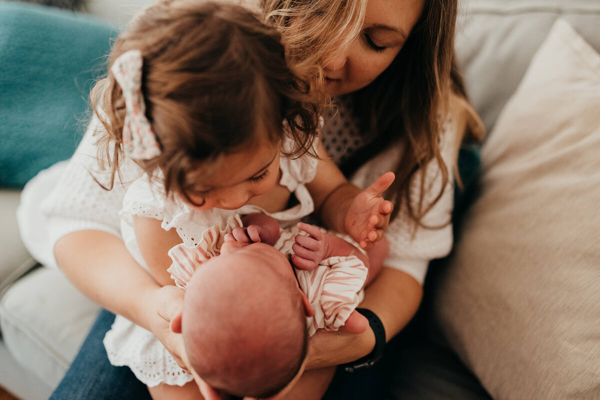 A mother holds her newborn daughter and her oldest daughter, the big sister is looking closely at the baby during their photoshoot with Allison Wolf, Boston Newborn Photographer