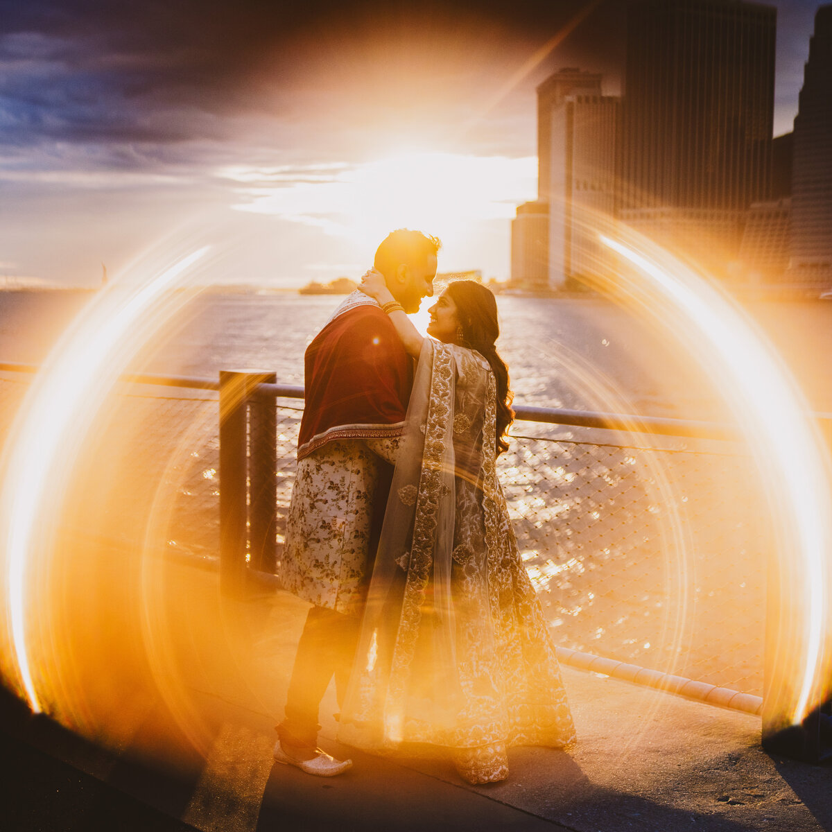 A couple share a moment in indian outfits at the promenade at brooklyn bridge park in dumbo with the ring of fire sunset