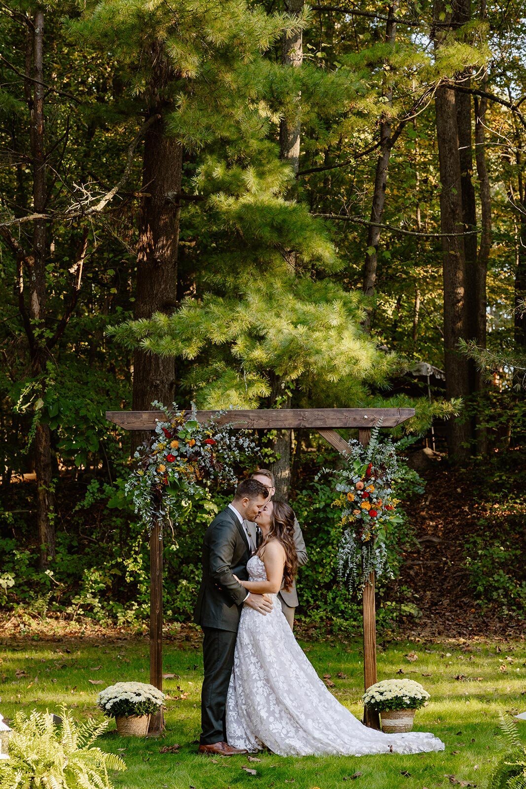 A bride and grooms first kiss on their wedding day. The couple is standing in front of a flower adorned arbor in the woods.