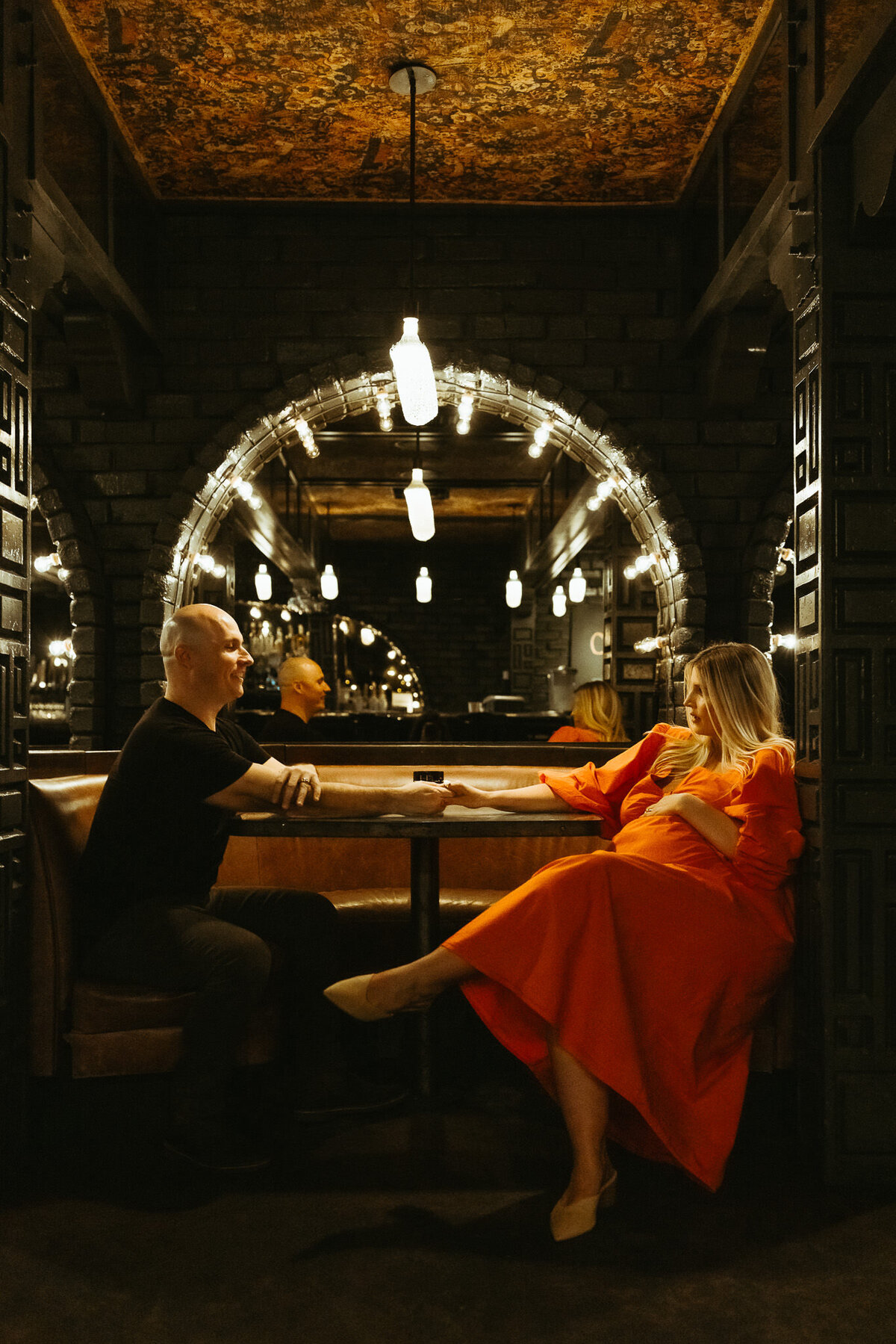 Maternity couple holds hands at table at Ace Hotel bar.  It is dark room with moody artificial lighting. Pregnant mom in orange dress and dad in black shirt.