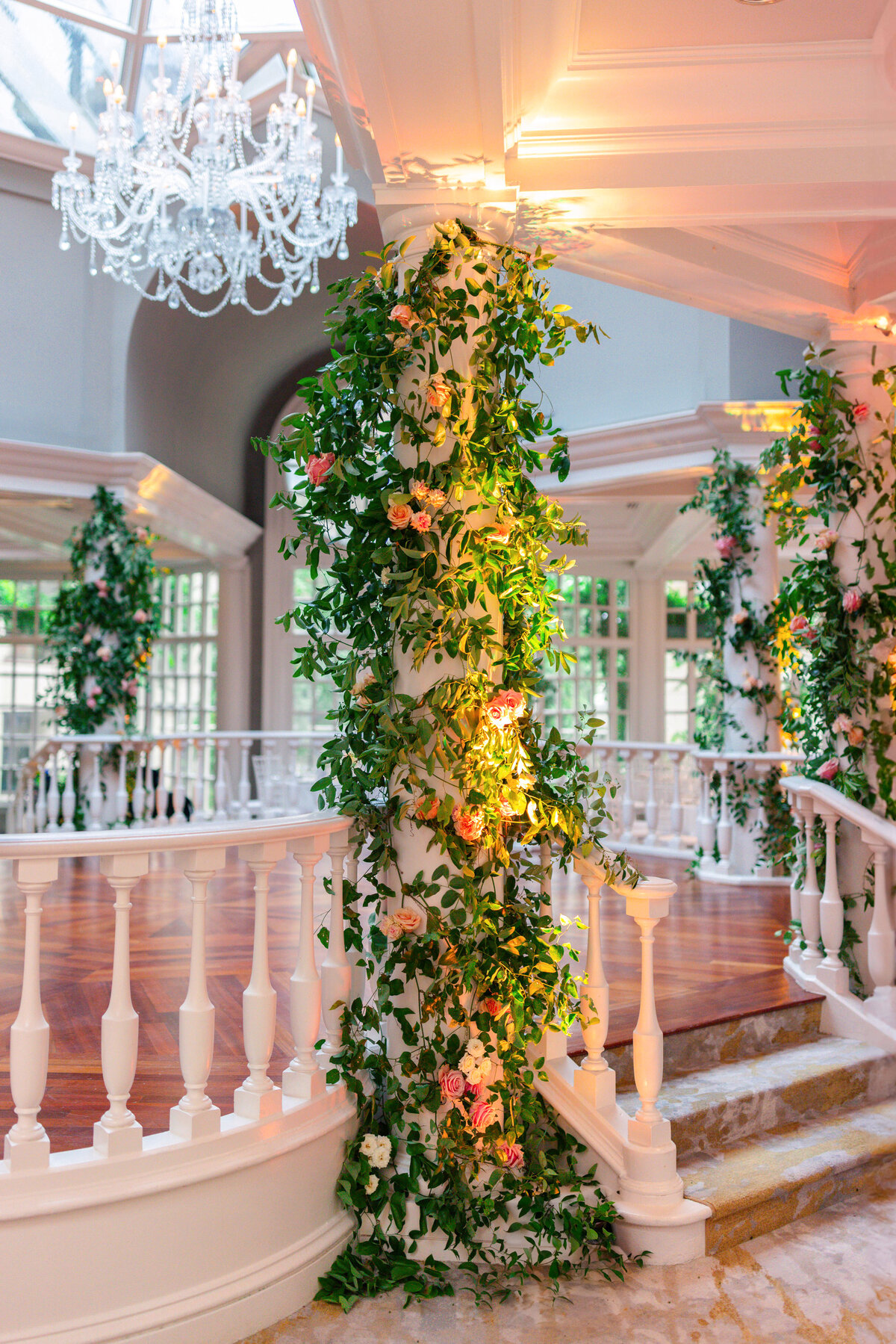 A white column wrapped in lush green vines and pink flowers stands in a sunlit room with a high ceiling. A nearby elegant chandelier hangs above, and the floor is wooden with white railings surrounding the area.