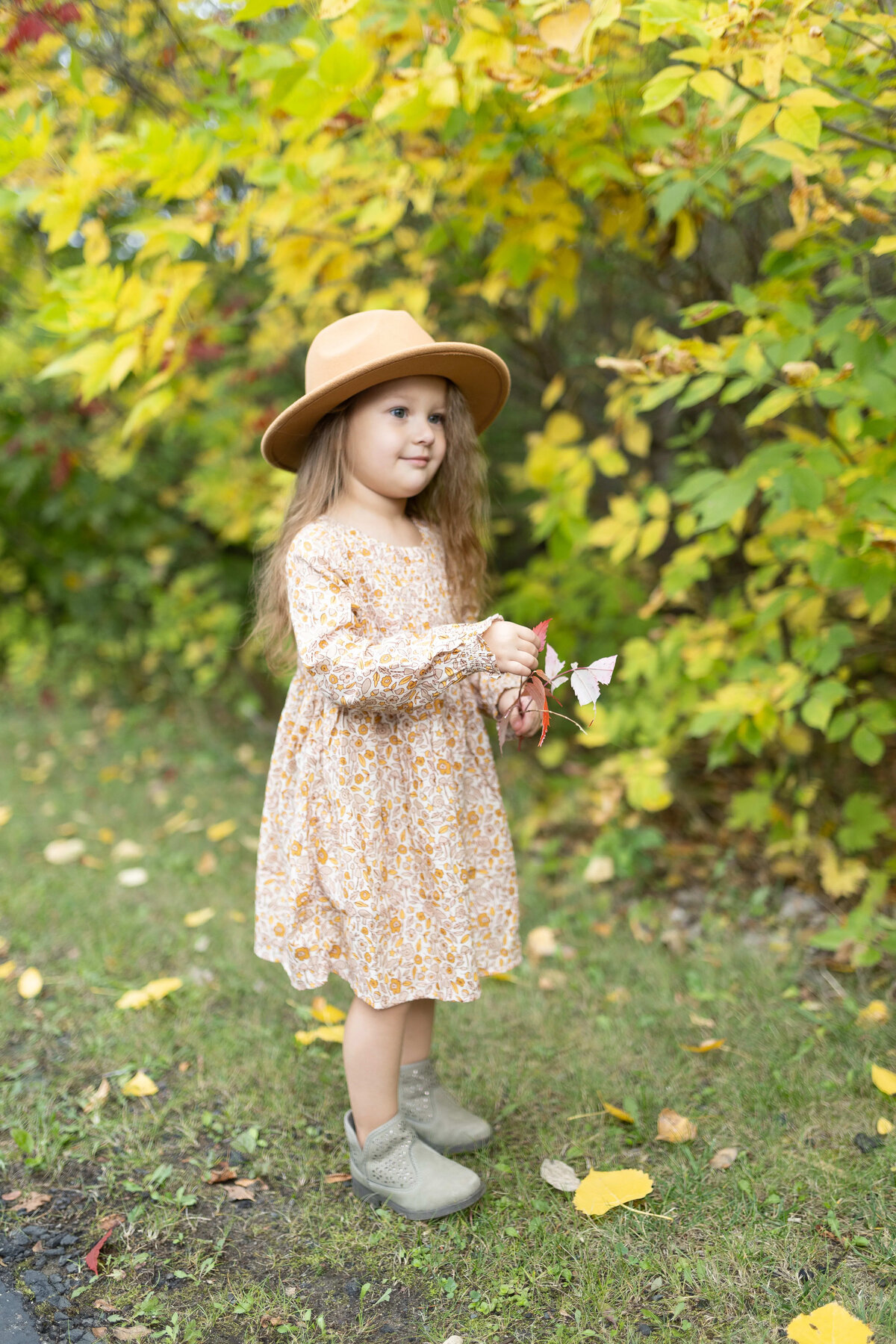 Little girl in mustard floral dress holding leaves in front of green foliage - outdoor children's portrait session