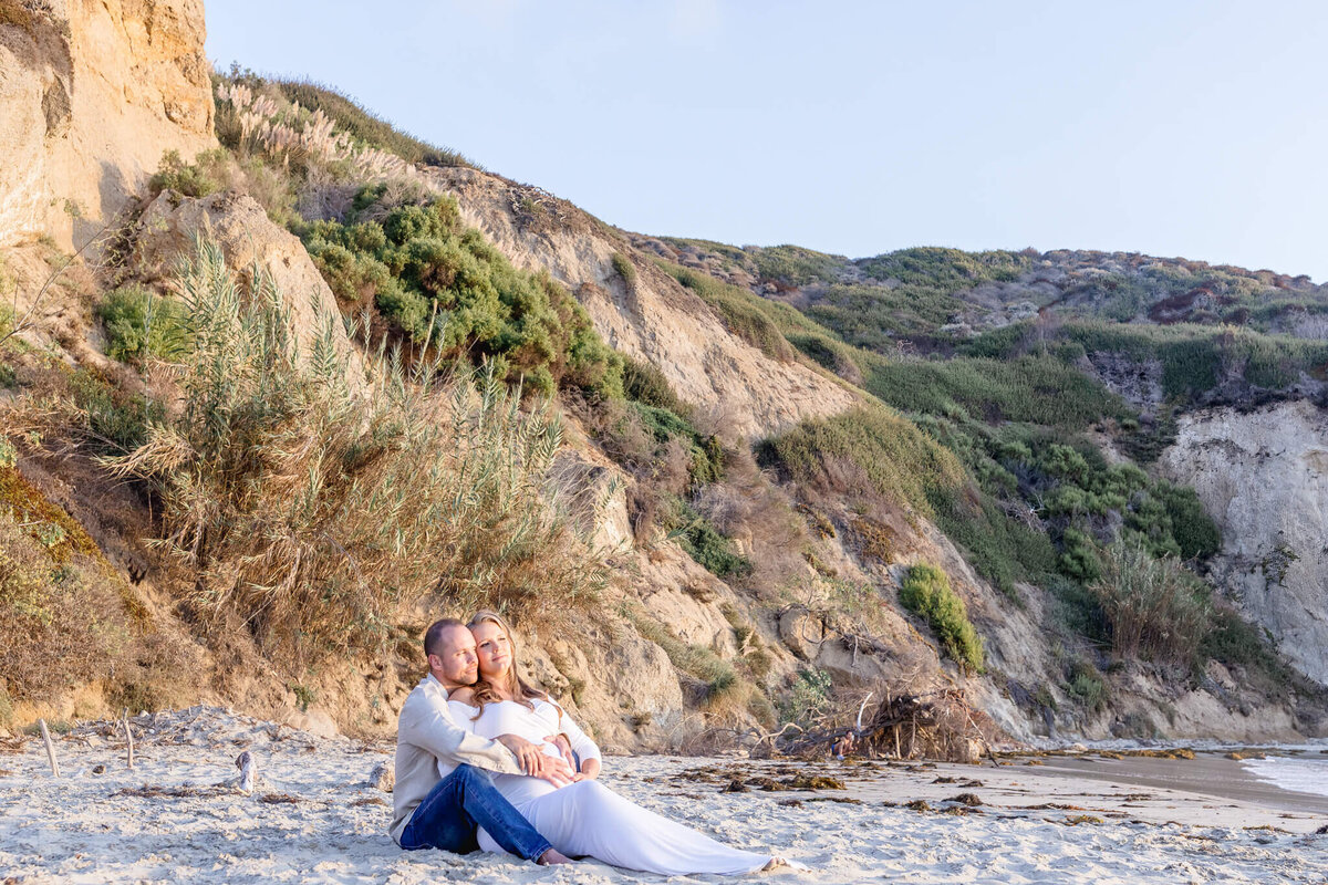 A couple sits together on a sandy beach, nestled against a backdrop of rocky cliffs covered in greenery. The sun casts a warm glow over the serene landscape.