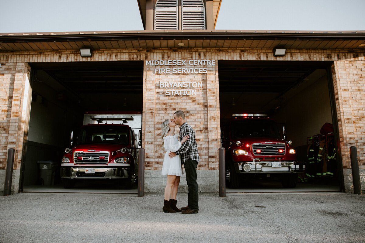 London, Ontario fire house engagement photoshoot. Man and woman are kissing in front of fire station. The bay doors are open and firetrucks are parked.