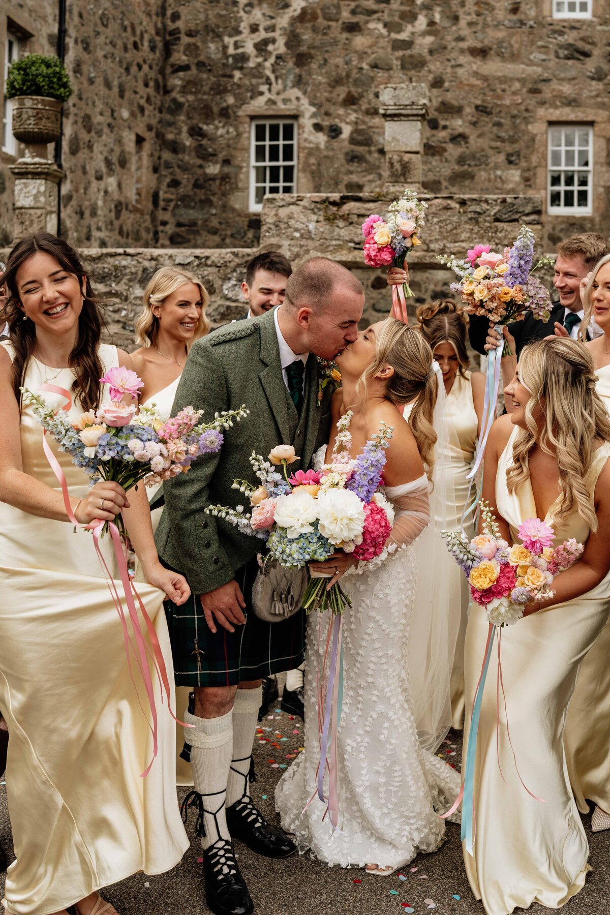 A bride and groom are photographed kissing as their wedding party jumps around in celebration around them. An Aberdeenshire castle, Barra Castle, is photographed behind them.