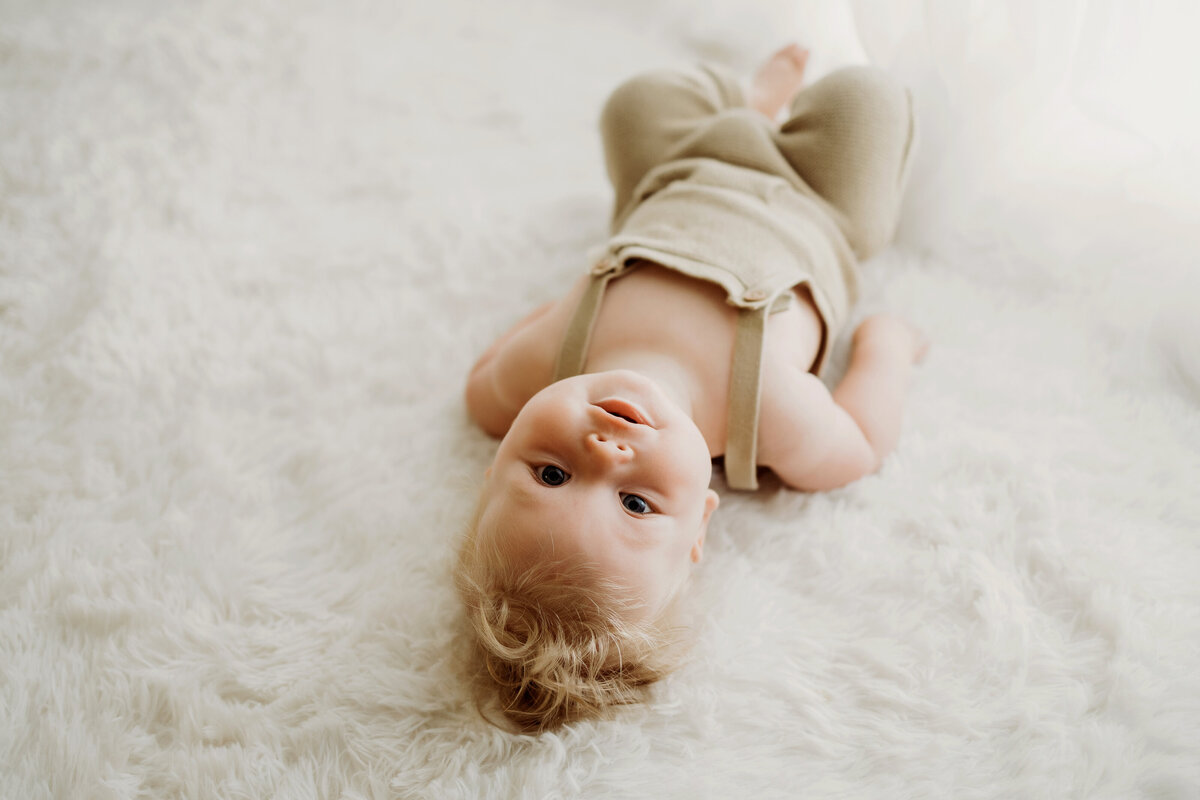 During his six month milestone session in Denver, a baby boy wearing a knit overalls lays on his back on a white rug