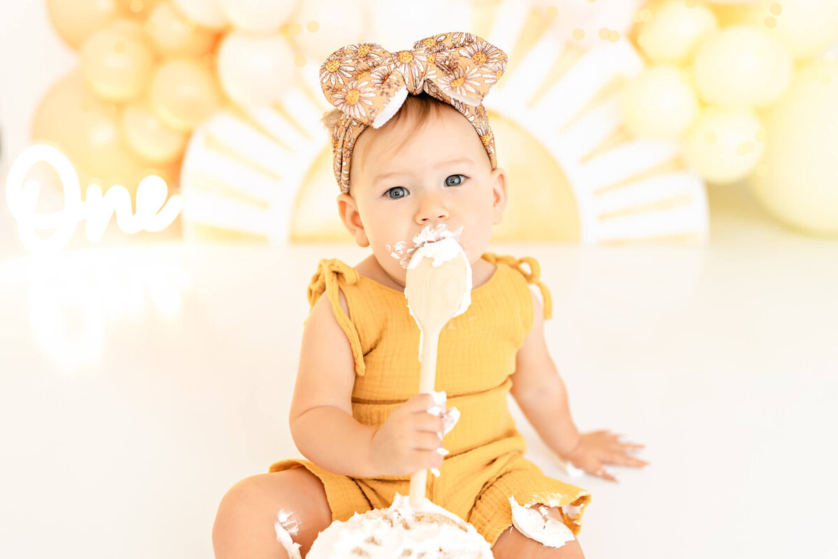 A baby wearing a mustard yellow outfit and a headband with a bow is sitting on the floor with a cake in front. The baby has cake on their face and holds a wooden spoon. There are yellow and white balloons and a "One" sign in the background.