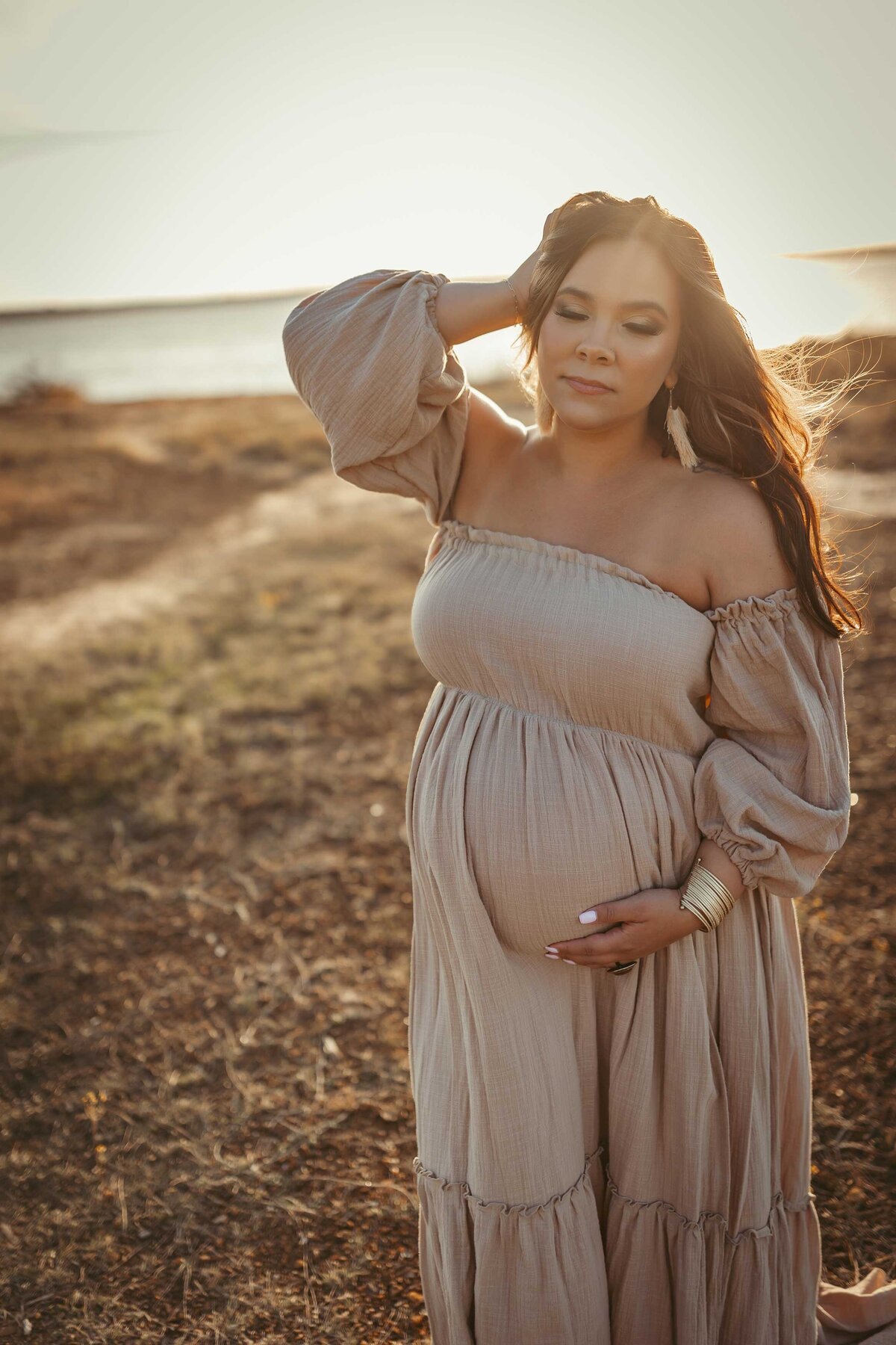 Pregnant woman standing close to the water. She is wearing a soft tan strapless dress and holding her hair with one of her hands.