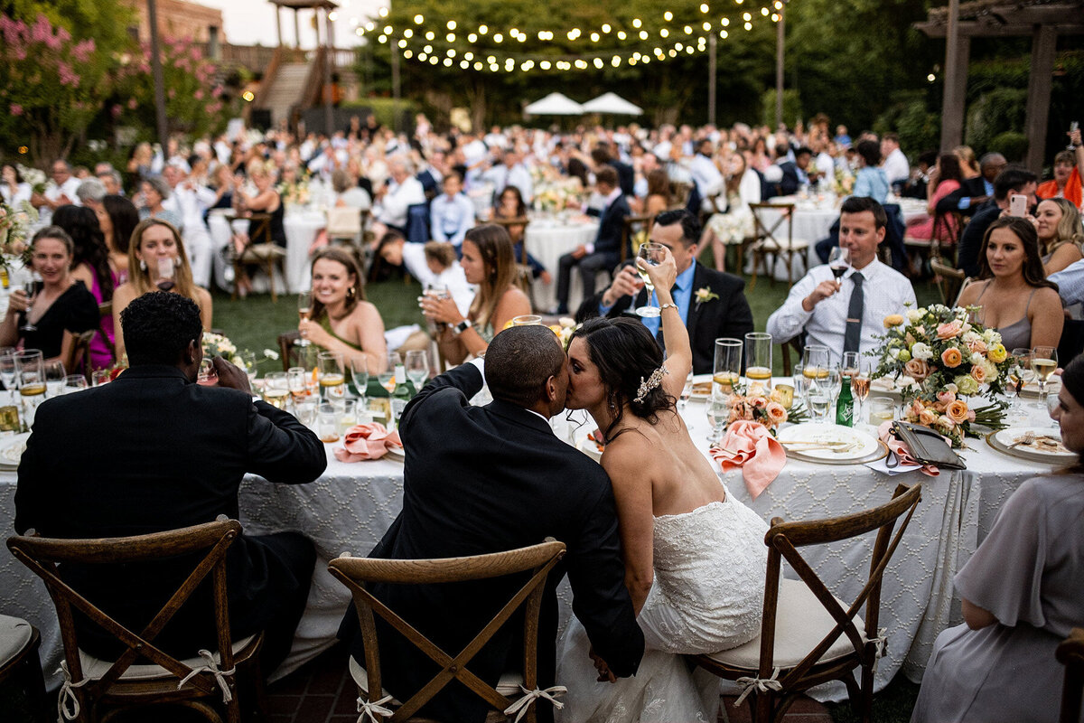 San Francisco Bay Area wedding photographer documents intimate bride and groom moment during outdoor reception dinner with celebrating guests and string lighting at Estate Yountville in Napa Valley