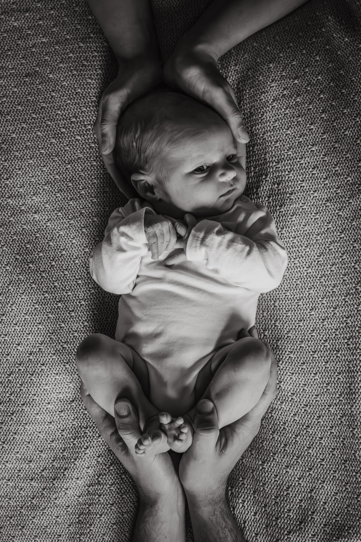 baby ligt op bed en papa houdt de beentjes vast en mama heeft de handen op het hoofdje.