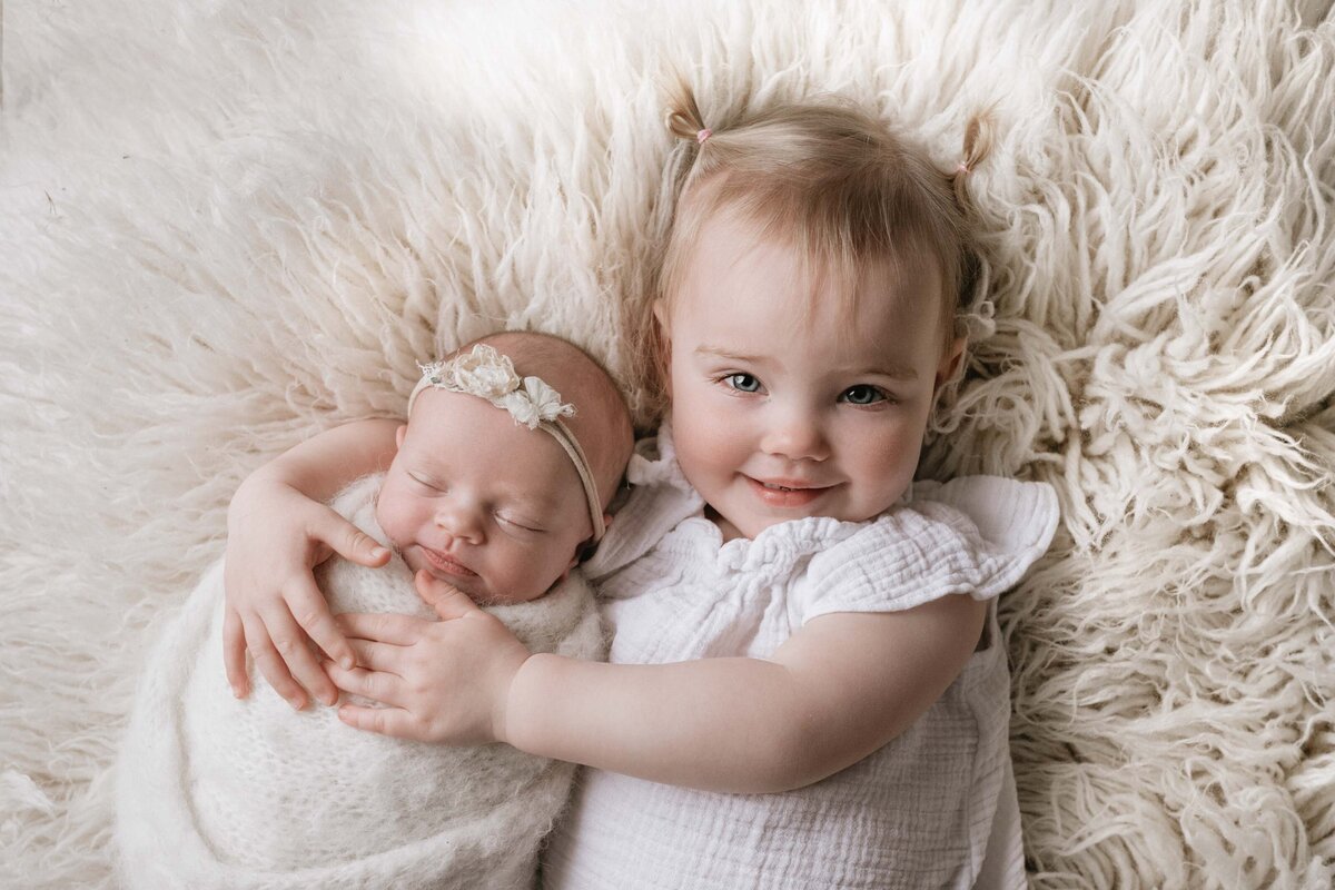 Toddler girl laying down holding newborn baby sister for posed newborn portraits. Baby girl and big sister are both matching in cream. Baby girl is sleeping and big sister has her arms draped around the sleeping baby and smiling at the camera.