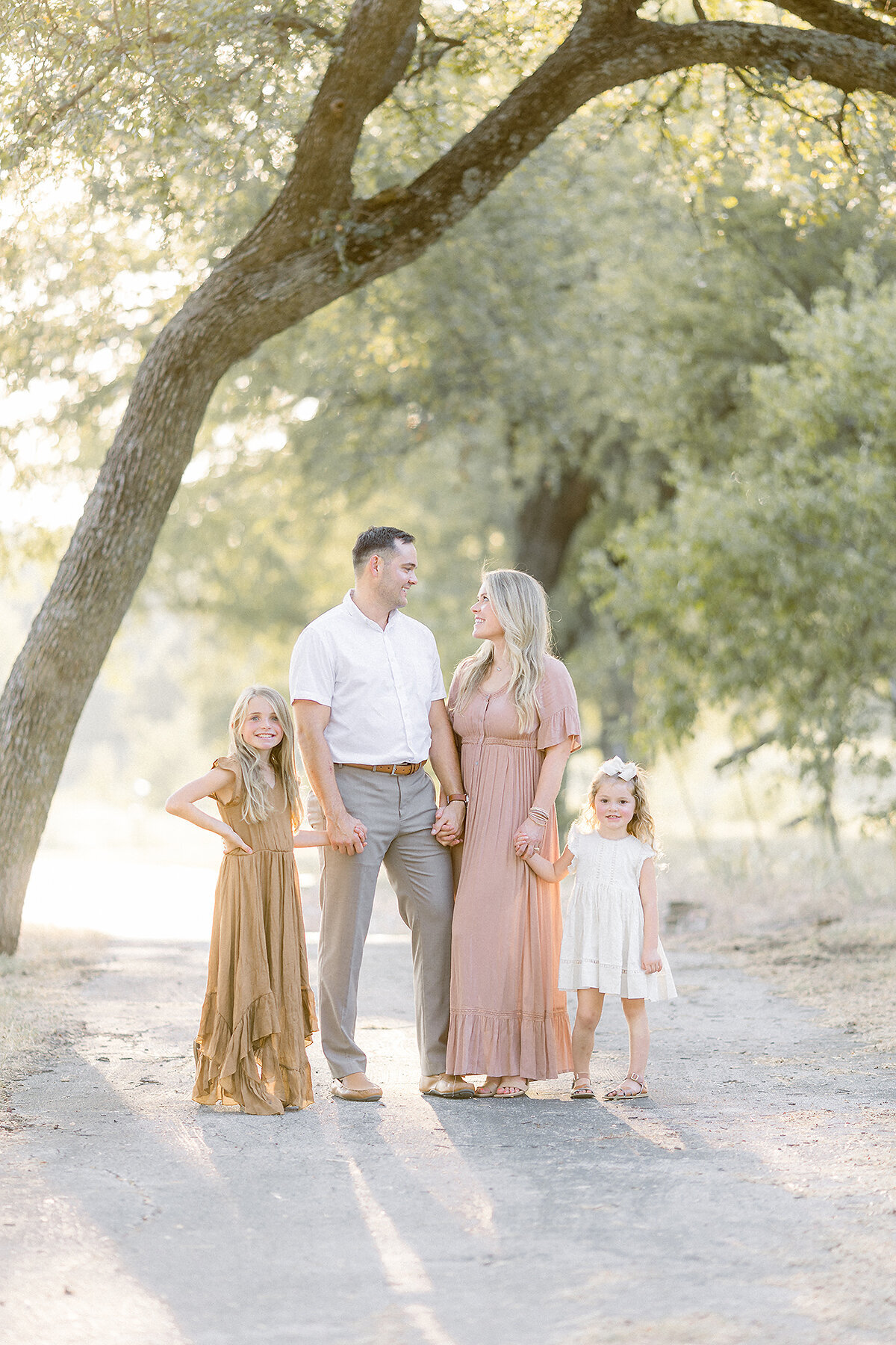 A young family of 4 portrait taken while they are all standing together in the middle of a path at a local Dallas/Fort Worth park as they look at each other while their family photographer captures this sweet moment.