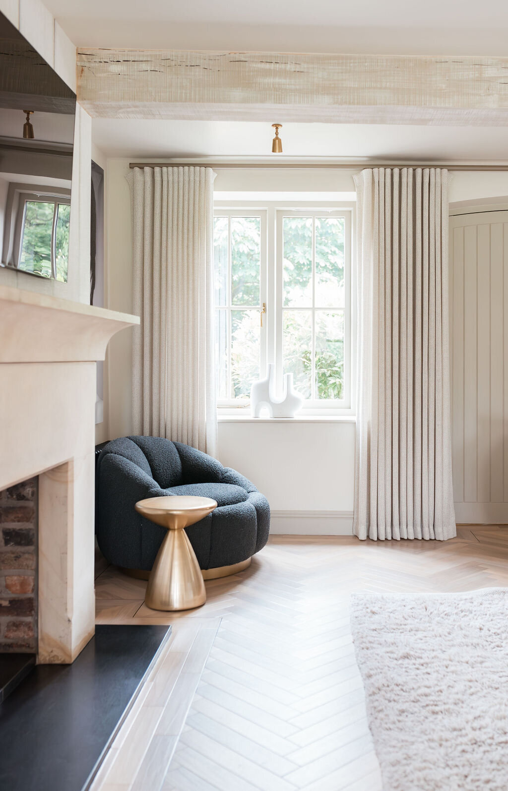 A cozy living room corner featuring a plush, dark gray armchair beside a small, round gold side table. The space is illuminated by natural light from the window draped with white curtains. The floor has light wood herringbone pattern, and the white walls create a calm ambiance.