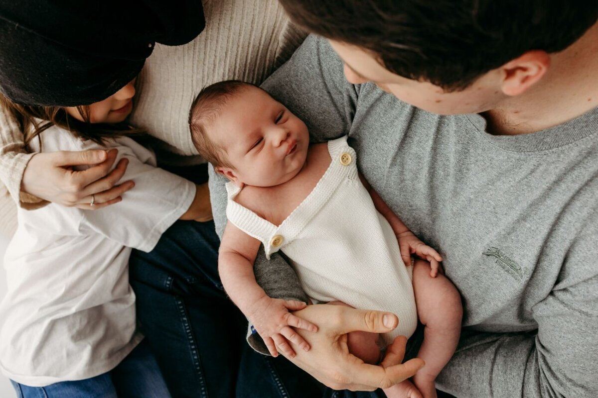 Top-down shot of a father holding his newborn baby, who is sleeping soundly, while the mother and an older child share a loving hug nearby.