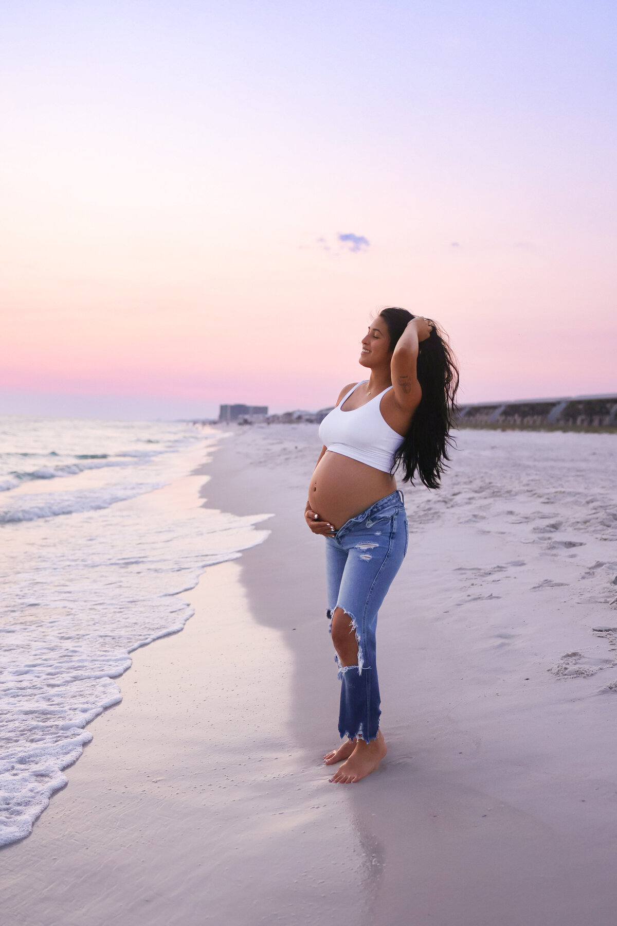 pregnant woman wearing white shirt and jeans standing on the beach at sunset in panama city beach florida