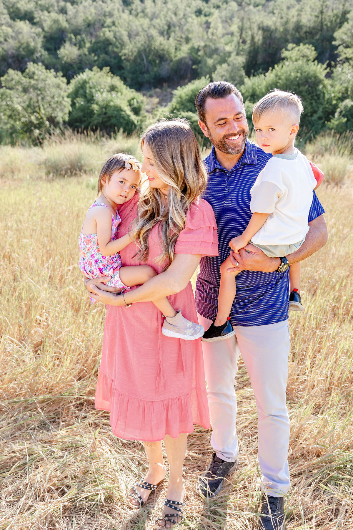 A young family stands closely together in a sunlit meadow. The mother in a pink dress holds her daughter while the father in a blue polo shirt carries their young son. Taken by Cassie Lee Photography