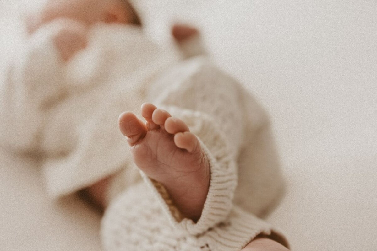 Detailed macro image of a newborn baby's small foot showing intricate wrinkles and tiny toes, clad in a cream knitted romper. Newborn photography