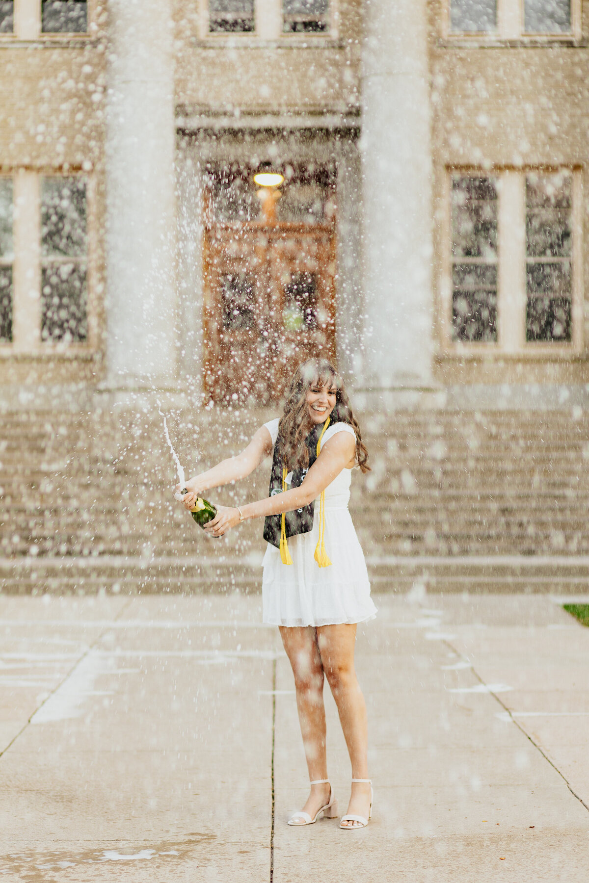 A Colorado State University graduate sprays a bottle of champagne in celebration during her grad session in Fort Collins, Colorado. The joyful moment captures her excitement and achievement, with the iconic CSU campus in the background.