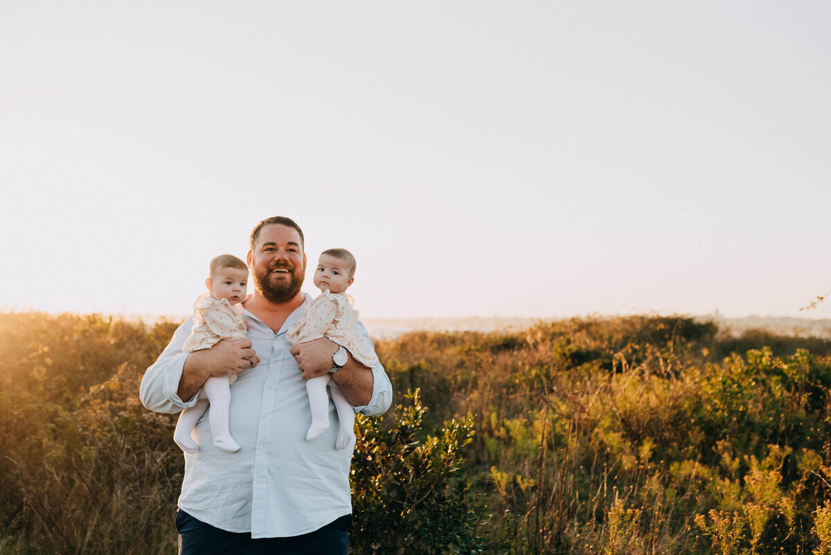 A father holds his twin baby girls in a field at sunset