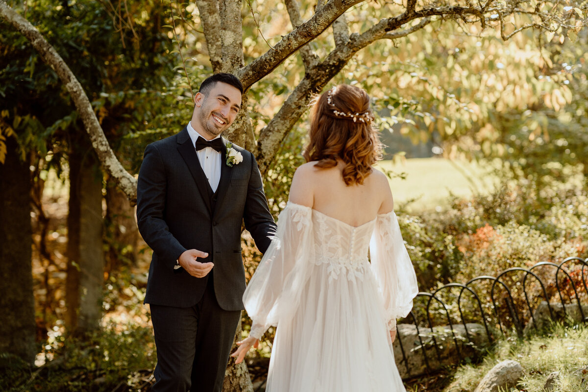 Groom smiles warmly as he sees his bride for the first time during their first look on their wedding day, set in a lush garden with trees and autumn foliage. The bride wears an off-the-shoulder gown with flowing sleeves, creating a romantic and enchanting atmosphere. Ideal for couples seeking a documentary-style wedding photographer to capture the joy and emotion of their wedding day