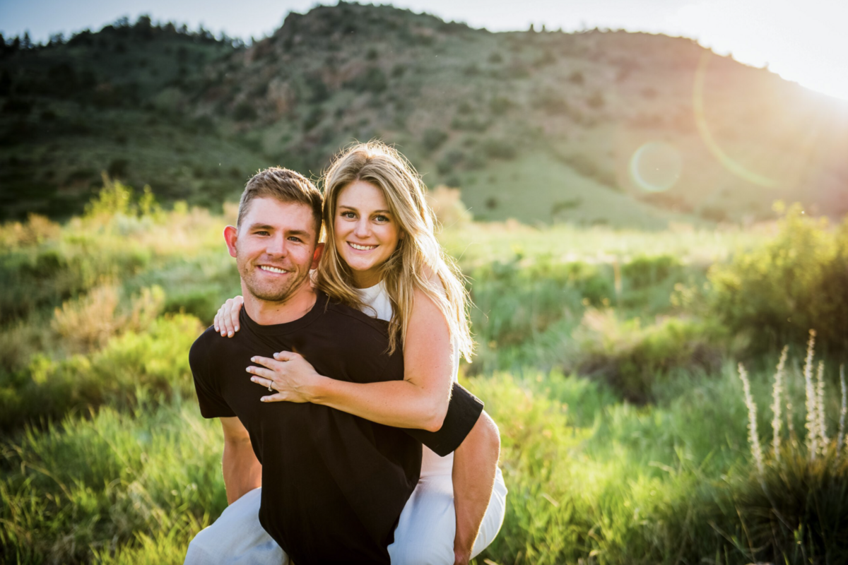 An engaged couple poses in a piggyback ride and smiles at the camera with a Colorado landscape in the background, captured by Denver engagement photographer, Casey Van Horn.