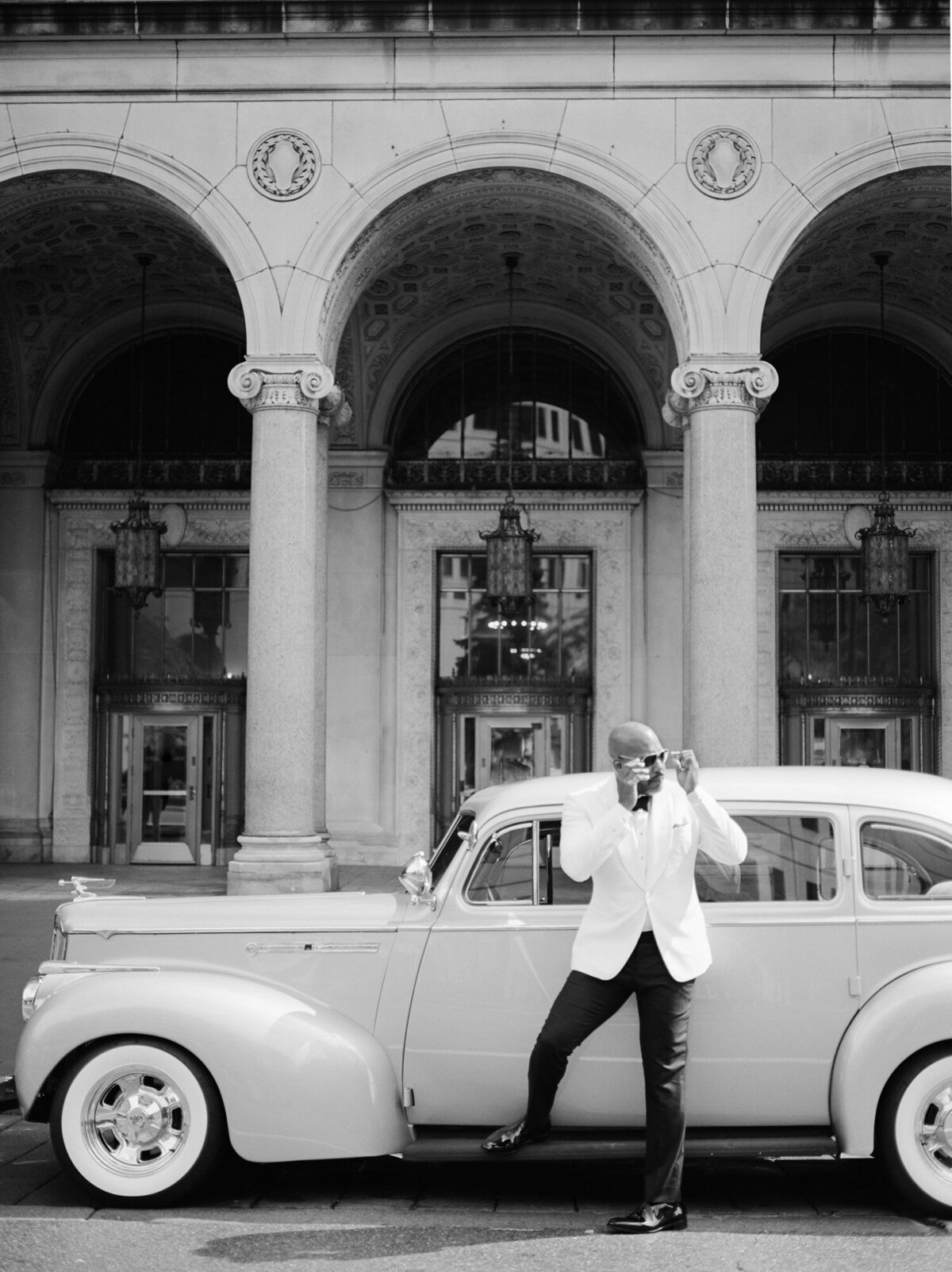 handsome black groom in white tuxedo jacket with sunglasses near vintage wedding car in Detroit, ornate architecture