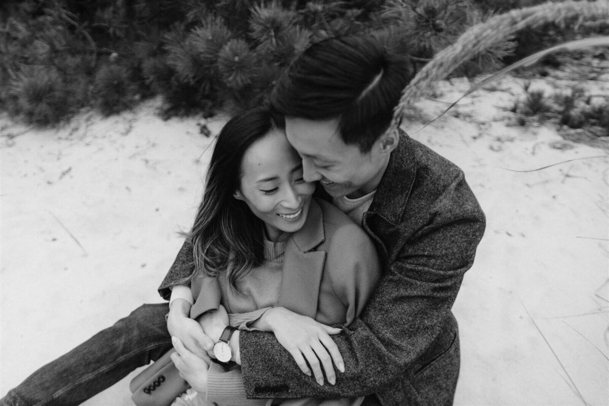 The man is hugging his fiancee while sitting on the white sand in Fire Island Beach, NY. Engagement Image by Jenny Fu Studio