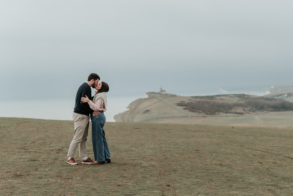 Newly engaged couple kiss in foreground with the South Downs landscape behind them
