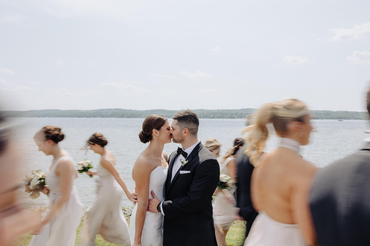 Bride and groom kissing whilst bridal party walking around them