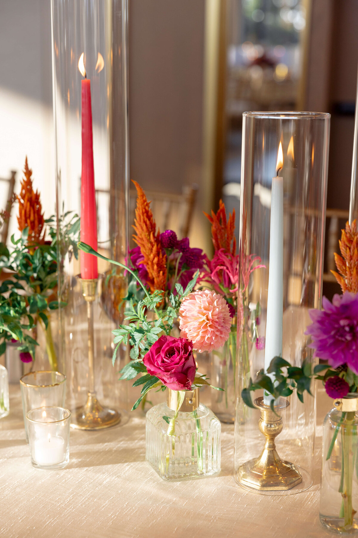 A table decorated with tall glass candle holders, one with a red candle and the other with a blue candle. Surrounded by vibrant flowers in shades of pink, purple, and orange, all set on a cream-colored tablecloth.
