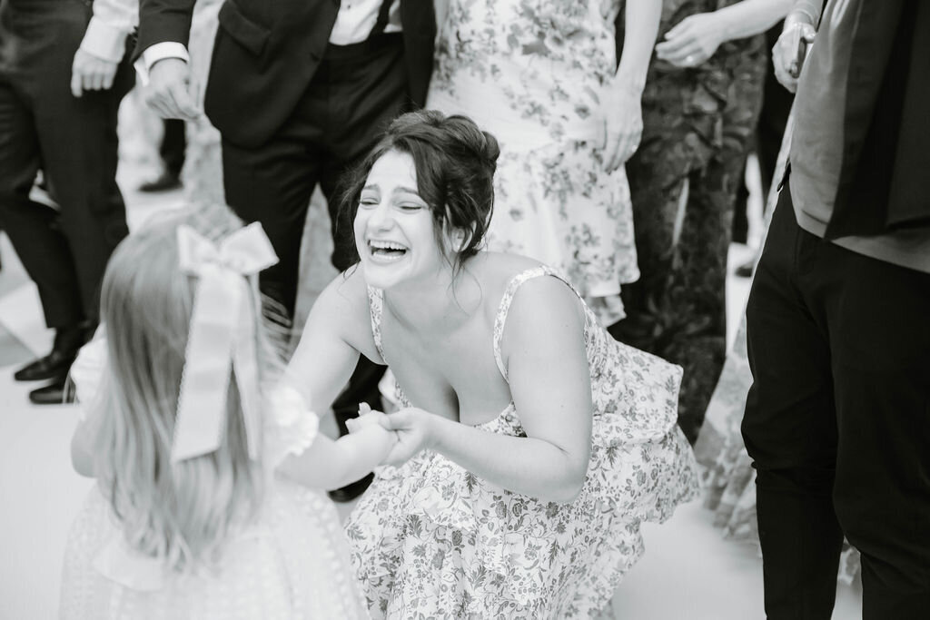 Rhode Island Wedding Photographers | A woman in a floral dress crouches joyfully, holding hands with a young girl in a dress with a large bow, surrounded by people in formal attire.