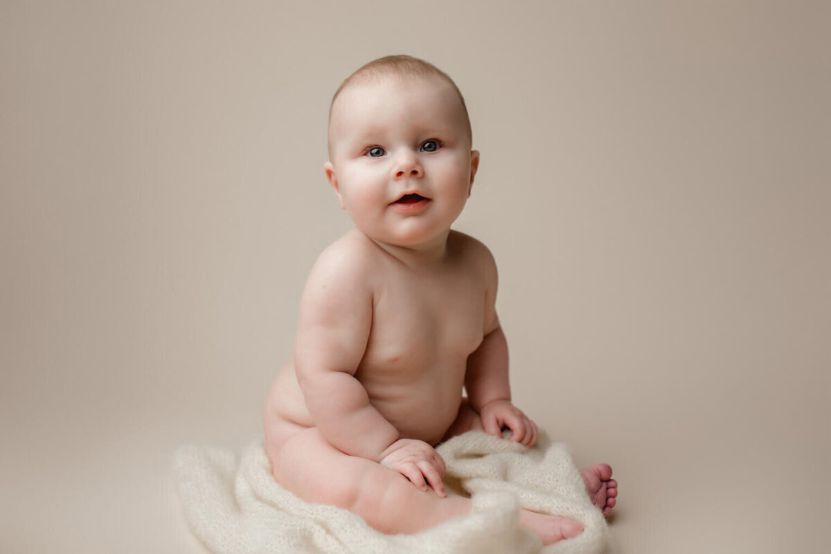 A chunky baby boy sitting on a white drop looking at the camera
