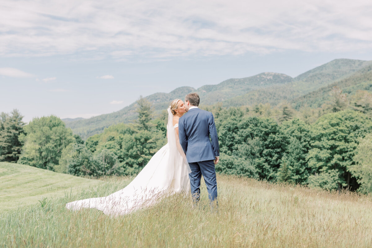 In front of the Adirondack mountains a bride and groom kiss in a tall grassy field on their wedding day in the middle of Summer.