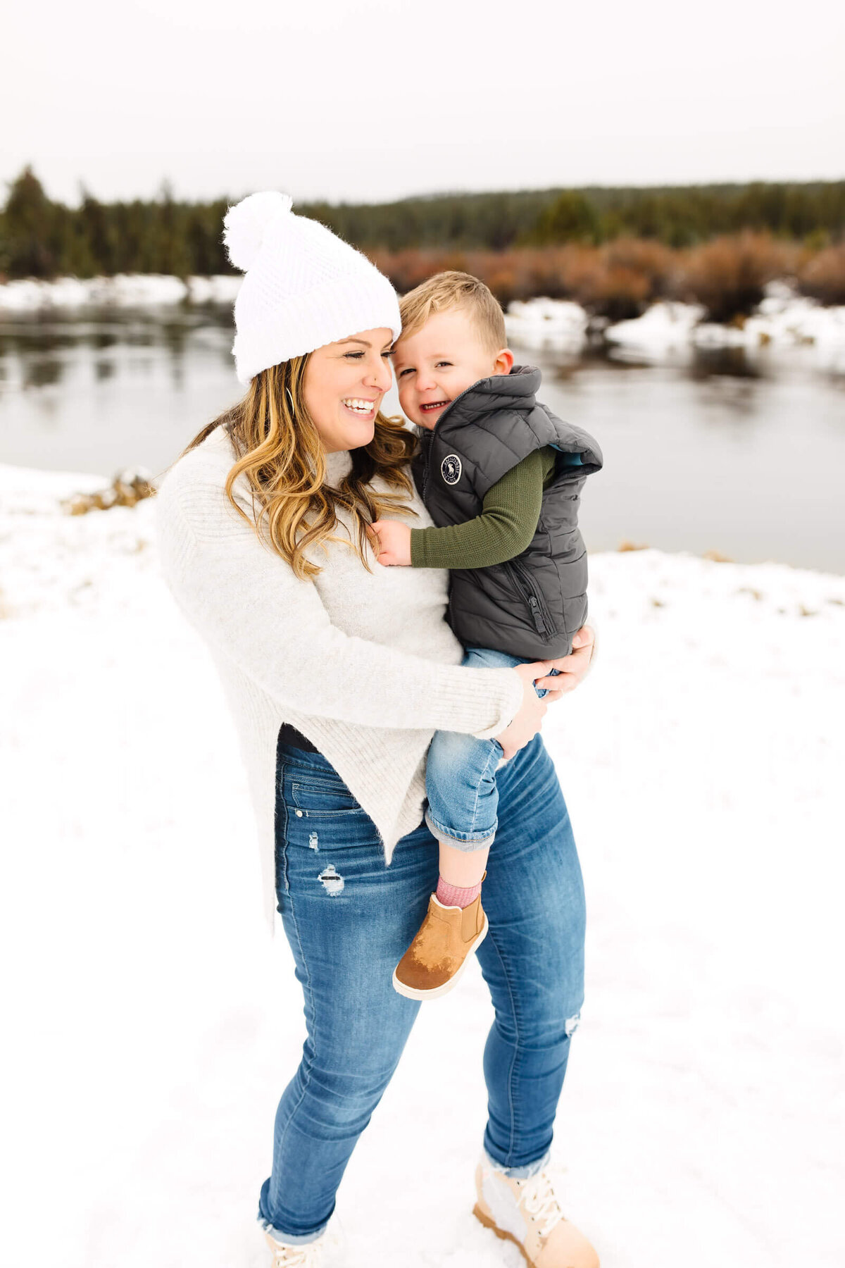 bend-family-photographer-mother-holding-baby-standing-in-snow-next-to-deschutes-river-1