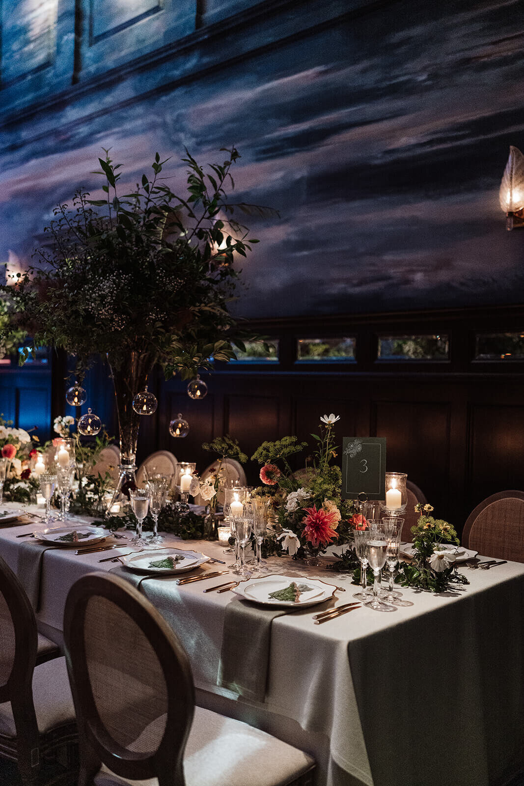 a long candlelit wedding dining table set with wicker round back chairs and trees as decoration in the nomad london ballroom for an intimate wedding