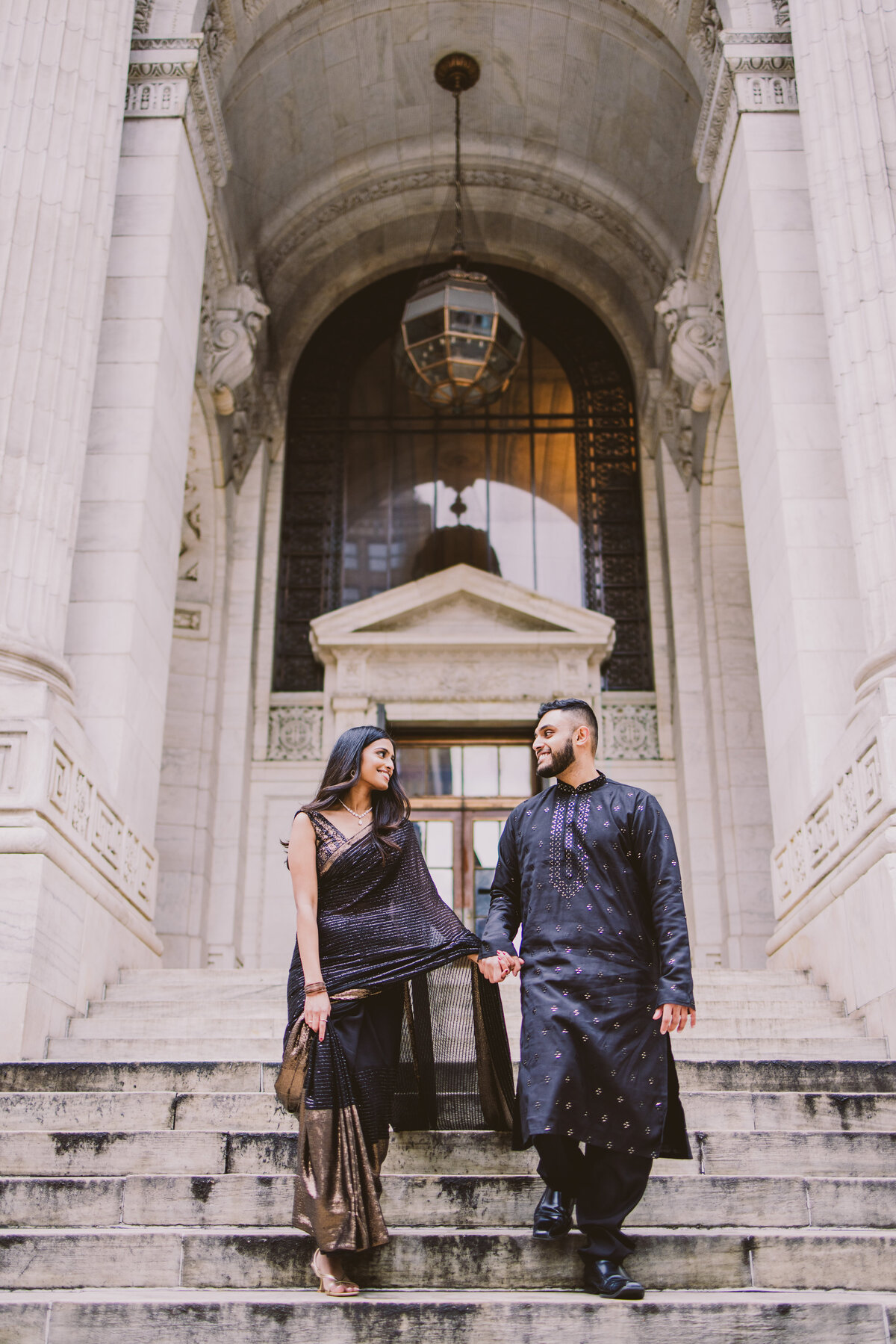 An engagement couple in traditional black indian outfits walking down the steps outside the new york public library in nyc