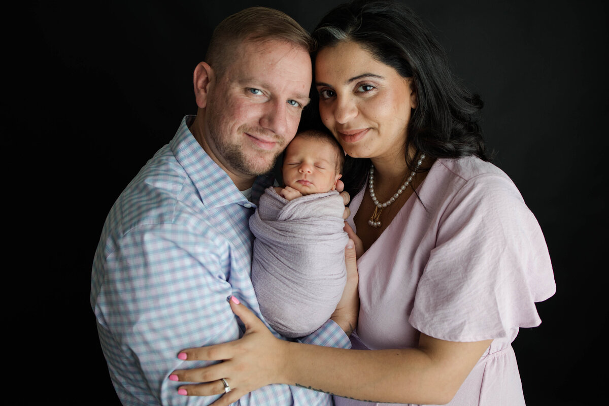 photo of Sleeping baby girl wrapped in a purple blanket being held in between mom and dad